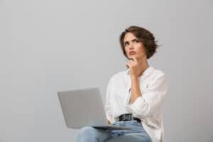 Thinking young woman sitting on stool isolated over grey background holding laptop computer.