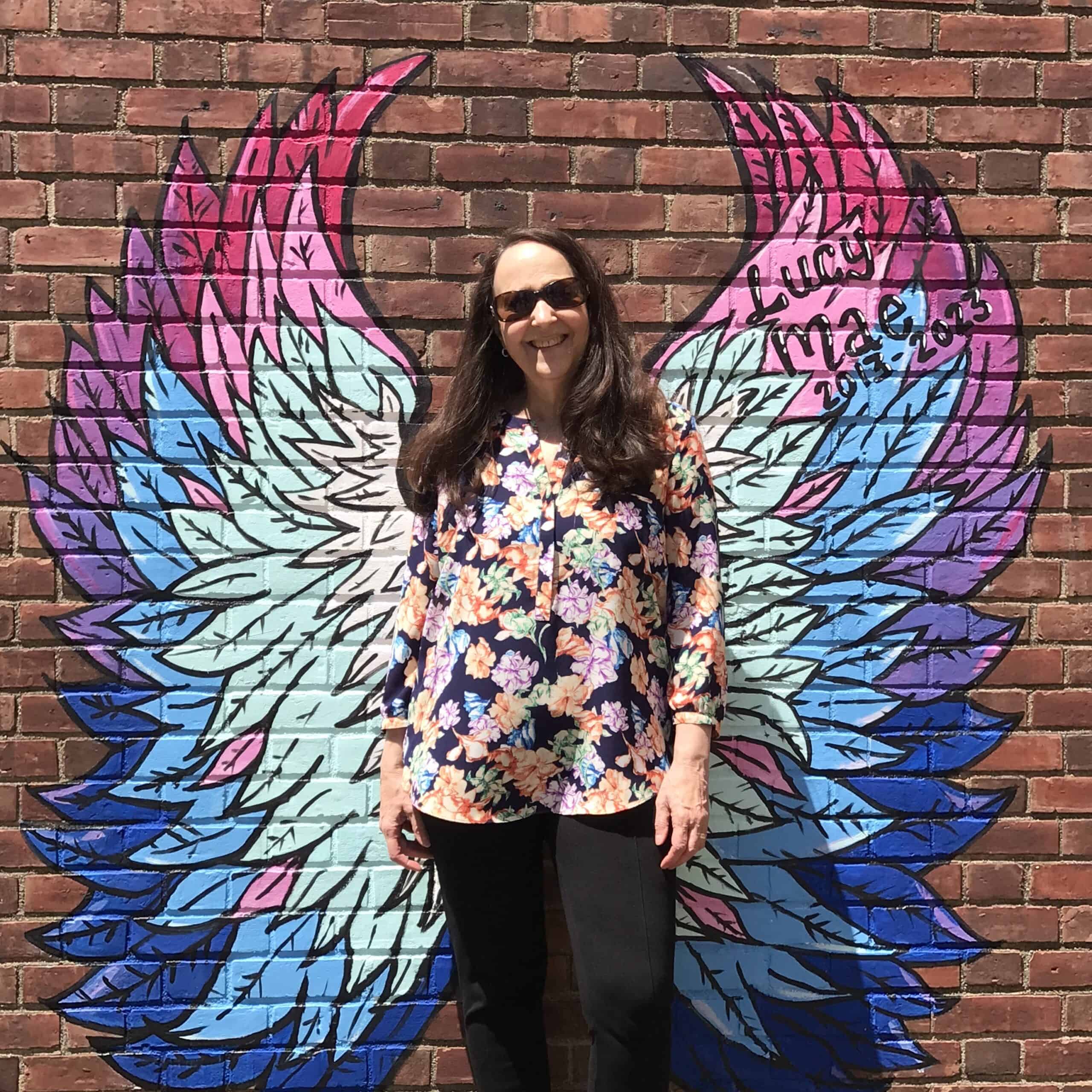 MelissaBrown-May2023 Melissa Brown in front of a brick wall with painted bright colored angel wings behind her