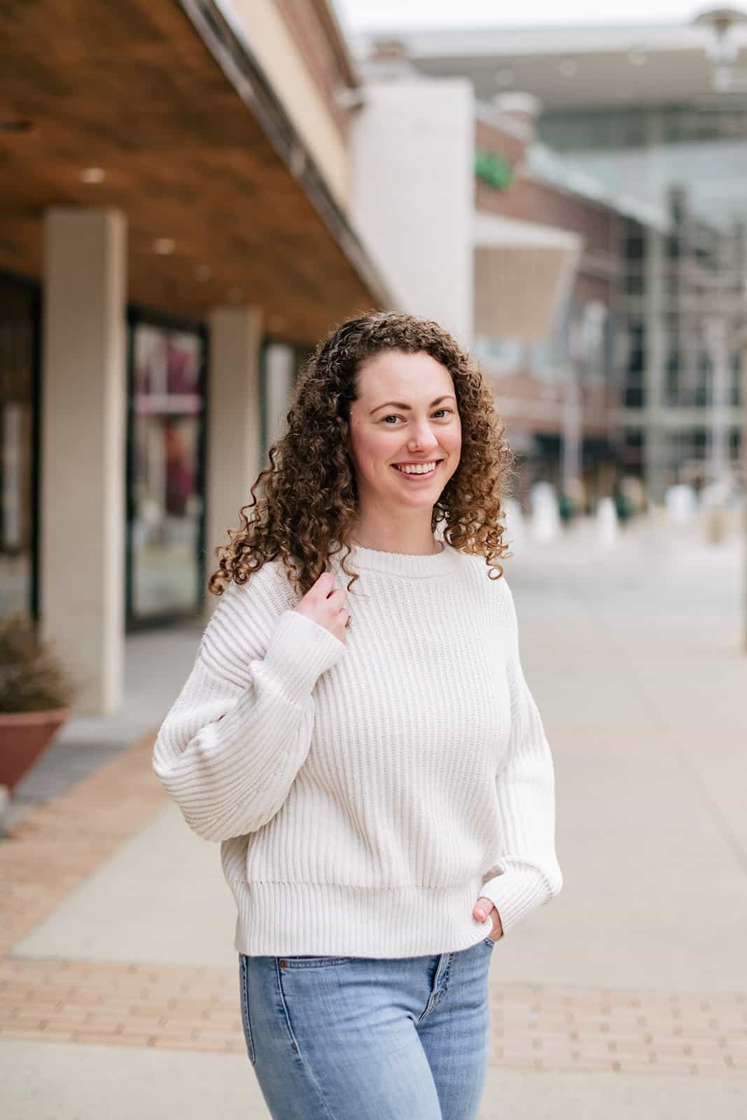 Brittany Herzberg A woman in a white sweater and jeans standing on a sidewalk.