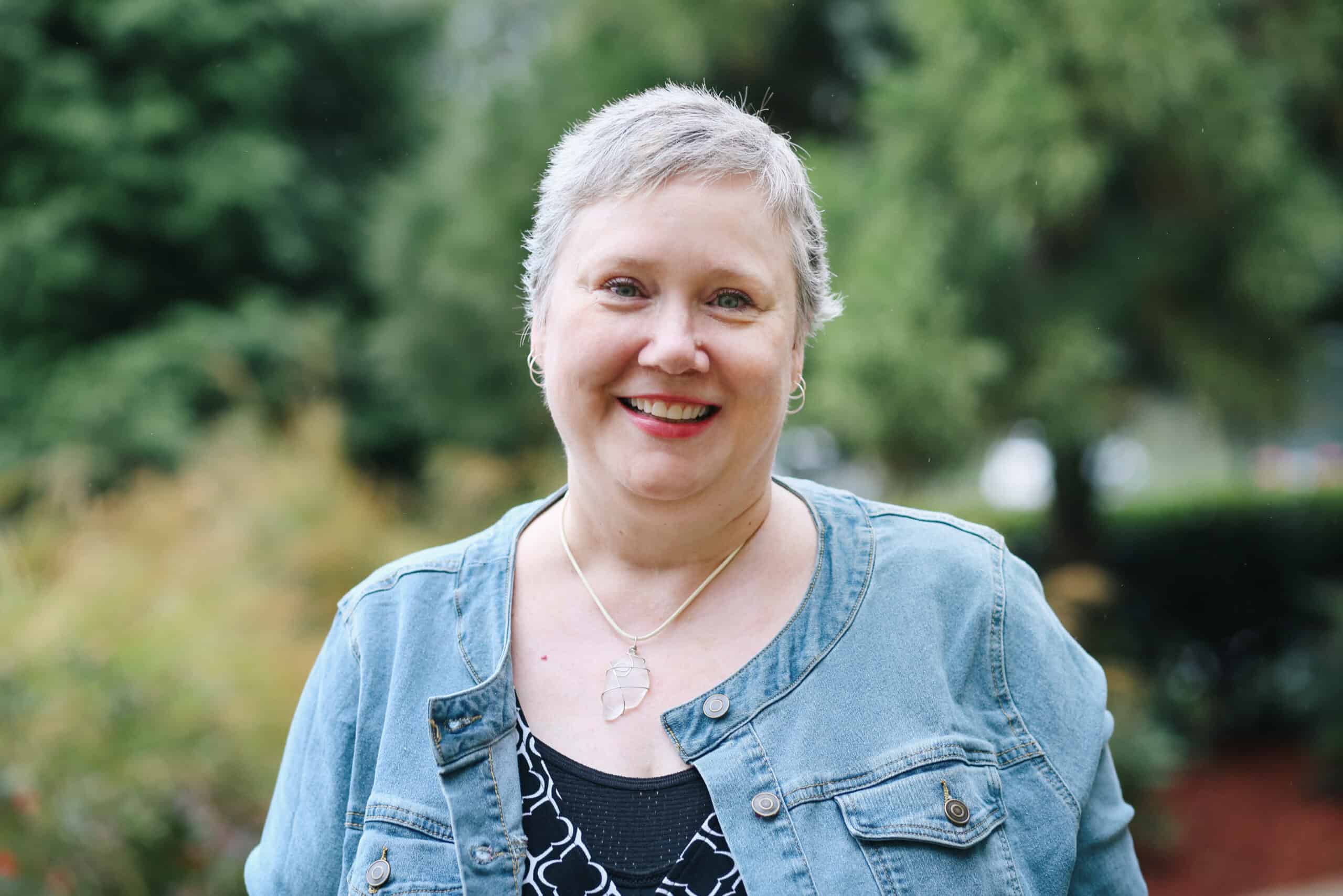 Kelly McCausey 2023 Kelly McCause in a denim jacket smiles in front of bushes.