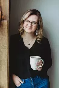 Woman smiling while holding a mug, leaning against a wooden door frame.