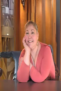 A smiling woman with short hair, wearing a coral top, resting her chin on her hand at a desk surrounded by wooden bookshelves.